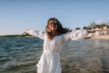 Photo, portrait of curly brunette, beautiful happy smiling young woman walking, walking in sunglasses on the water in the sea, ocean on the beach at sunset in a white sundress, dress.