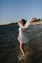 Photo, portrait of a beautiful happy smiling young woman walking, walking in sunglasses on the water in the sea, ocean on the beach at sunset in a white sundress, dress.