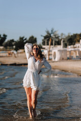 Photo, portrait of a beautiful happy smiling young woman walking, walking in sunglasses on the water in the sea, ocean on the beach at sunset in a white sundress, dress.