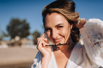 Photo, close-up portrait of curly beautiful happy smiling young woman model in sunglasses on the beach against the backdrop of the sea, ocean in a white sundress, dress at sunset.