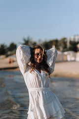 Photo, portrait of a beautiful happy smiling young woman walking, walking in sunglasses on the water in the sea, ocean on the beach of the island at sunset in a white sundress, dress.