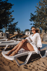 Curly brunette, beautiful happy smiling young woman in sunglasses lies, sits on a sun lounger on the beach in the sand in a white sundress, dress. Photography, portrait.