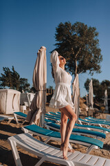 Curly beautiful happy smiling young woman model in sunglasses standing on sunbed on sandy beach in white sarafan, dress relaxing on vacation. Photography, portrait.