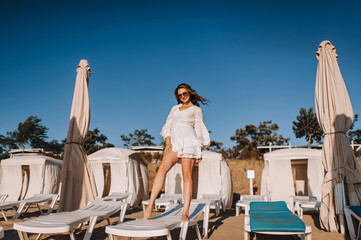 Curly beautiful happy smiling young woman model in sunglasses standing on sunbed on sandy beach in white sarafan, dress relaxing on vacation in summer. Photography, portrait.