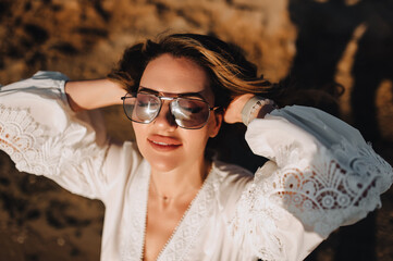 Photo, close-up portrait of curly beautiful happy smiling young woman model in sunglasses on the beach against the backdrop of the sea, ocean in a white sundress, dress at sunset.