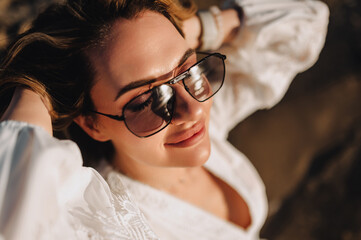 Photo, portrait of curly beautiful happy smiling young woman model in sunglasses during the day on the beach at sea, ocean in a white sundress, dress at sunset.