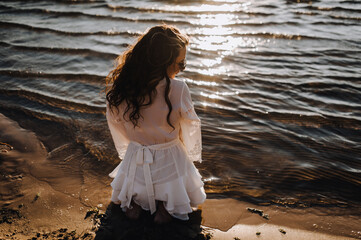 Photograph, portrait of a beautiful curly woman sitting on the sand on the beach against the backdrop of the sea, ocean in a white sundress, dress at sunset.