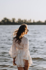 Photo, portrait of curly brunette, beautiful happy smiling young woman walking, walking in sunglasses on the water in the sea, ocean on the beach at sunset in a white sundress, dress.