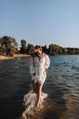Photo, portrait of a beautiful happy smiling young woman walking, walking in sunglasses on the water in the sea, ocean on the beach at sunset in a white sundress, dress.