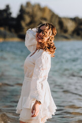 Photo, portrait of curly brunette, beautiful happy smiling young woman walking, walking in sunglasses on the water in the sea, ocean on the beach at sunset in a white sundress, dress.