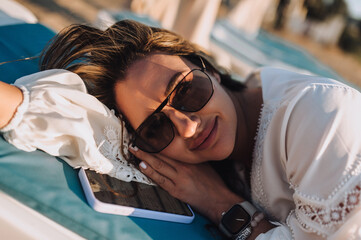 Curly happy smiling young woman in sunglasses lies with phone on sunbed in white sarafan, dress at sunset. Photography, portrait.