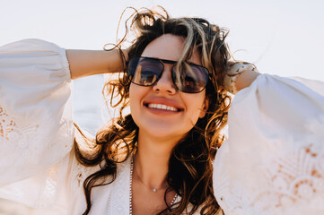 Photo, close-up portrait of a curly beautiful happy smiling cheerful young woman model in sunglasses on the beach against the backdrop of the sea, ocean in a white sundress, dress at sunset.