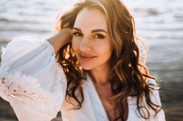 Photo, close-up portrait of curly beautiful happy smiling young woman model in sunglasses on the beach against the backdrop of the sea, ocean in a white sundress, dress at sunset.