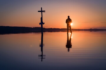 Silhouette of a person standing in water with a cross during sunset and reflection on water
