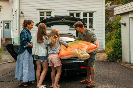 Daughters helping father loading pool raft in car trunk at driveway for road trip