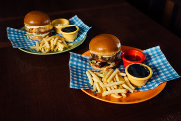 Two Oktoberfest burgers with tomato, cheese, and sauce, served with French fries and sauces. Presented on a plate with a Bavarian checkered napkin (lozenge pattern), evoking the spirit of Oktoberfest.