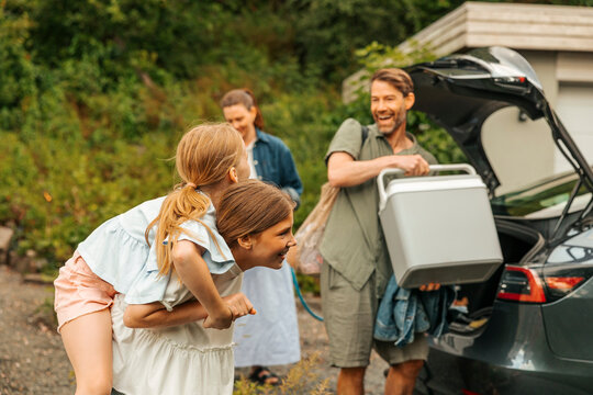 Happy man loading cooler in car trunk while looking at daughter piggybacking sister