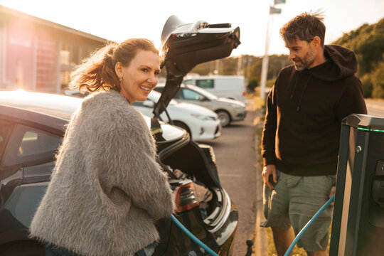 Smiling woman refueling electric car at charging station on street