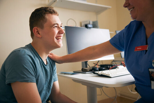 Happy female healthcare expert talking with hand on shoulder of young man in doctor's office