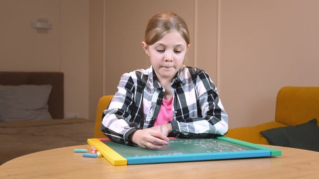 Primary school student practicing drawing on a small blackboard placed on a round wooden table near a mustard-colored sofa in a cozy living room in an apartment