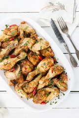 Succulent chicken wings and drumsticks are piled high on a white platter, garnished with herbs and vibrant red peppers, on a white wooden table. Utensils await