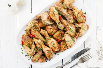 A delicious pile of cooked chicken pieces, including wings and drumsticks, sits on a white serving tray. The dish is garnished with fresh herbs and red chili peppers for added zing
