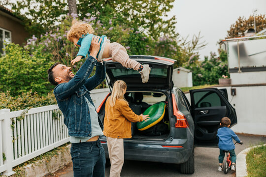Father playing with daughter while woman loading luggage in car trunk