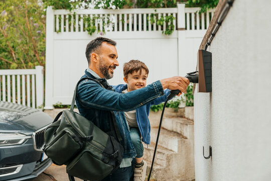 Smiling man carrying son while plugging charger in electric socket on wall
