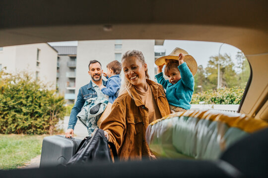 Happy woman holding daughter and loading bags in car trunk for weekend trip