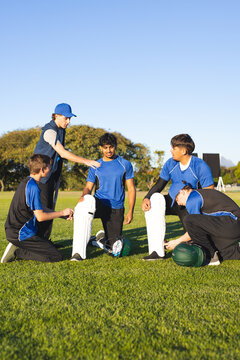 Diverse male cricket team in blue jerseys tying leg pads by stumps, helmets on cricket field