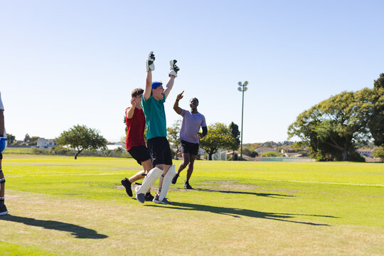 Diverse male teammates celebrating cricket play on field in wicket-keeping gloves and leg pads - Powered by Adobe