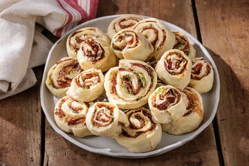 Plate heaped with freshly baked savory pinwheels, showcasing layers of dough, pesto, and cured meat. Served on a rustic wooden table, promising a delicious snack