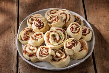 A plate overflows with freshly baked pinwheels. Sun-dried tomatoes and herbs nestle in flaky pastry, creating appetizing swirls. The Rustic wood board below completes the scene