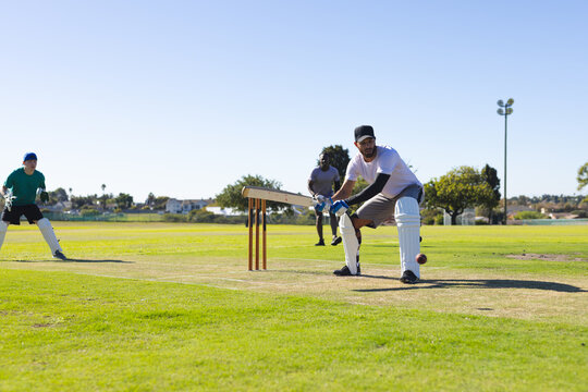 Diverse male players playing cricket on grassy pitch with bat, red ball, stumps, copy space - Powered by Adobe