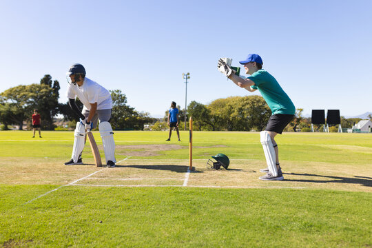 Diverse male teammates practicing batting at crease on cricket pitch with bat near wicket stumps