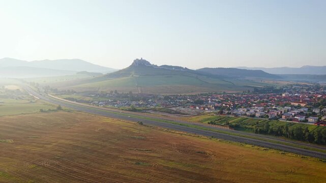 Aerial view of fields and the D1 highway near Spi&scaron;sk&eacute; Podhradie, Slovakia, with a quaint town nestled among rolling hills under a clear sky.