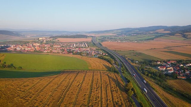 Aerial view of fields and the D1 highway near Spi&scaron;sk&eacute; Podhradie, Slovakia, with a quaint town nestled among rolling hills under a clear sky.
