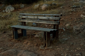 Old wooden bench in a rustic outdoor setting, standing on rocky dry ground with grass and stones around. Weathered and abandoned seat in nature, symbolizing solitude, nostalgia, and simplicity of rura