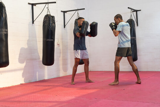 African american male trainer with trainee in sportswear practicing boxing in gym with focus mitts