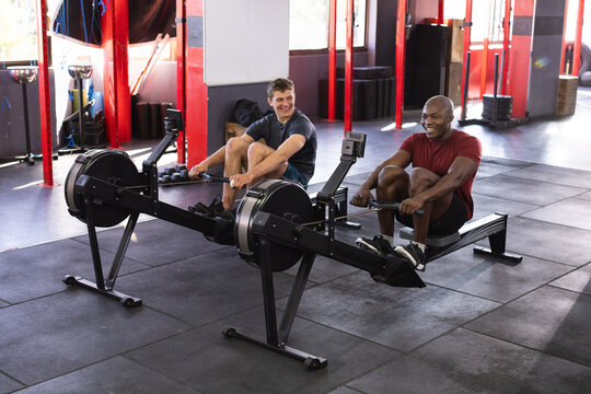 Diverse male friends rowing on rowing machines in gym surrounded by weight plates, dumbbell rack