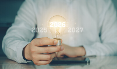 Hand holding glowing light bulb with futuristic overlay showing year 2026, symbolizing ideas, innovation, and planning for the future.