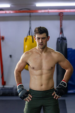 Man in 30s standing arms akimbo in fingerless gloves in boxing gym beside punching bags