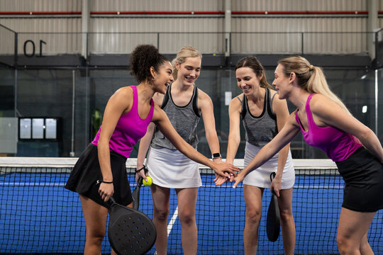 Diverse female teammates in sportswear holding padel rackets stacking hands on net on court - Powered by Adobe