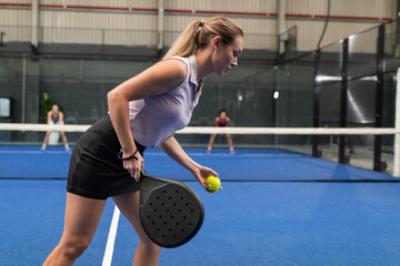 Diverse female opponents preparing serve with racket, ball over net on blue padel court