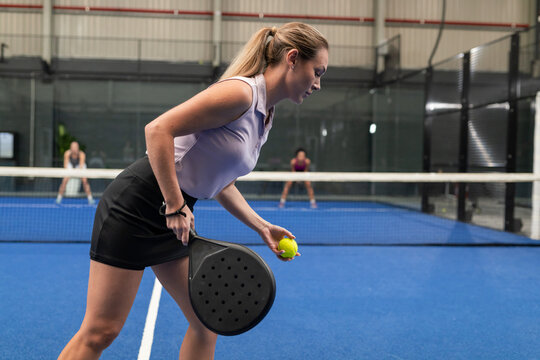 Diverse female opponents preparing serve with racket, ball over net on blue padel court