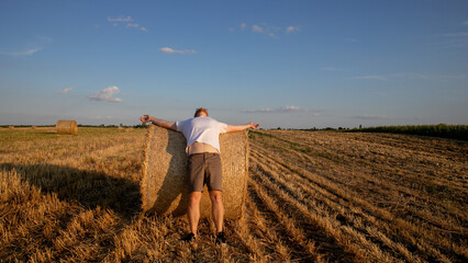 A Caucasian man stretches like a modern-day scarecrow against vibrant twilight hay bales, evoking...