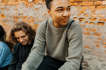Young man looking away while sitting with friends in front of brick wall