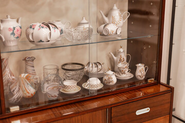A wooden display cabinet contains an array of vintage porcelain teapots, cups, and glassware. The intricate designs highlight skilled craftsmanship and a passion for decorative arts