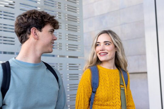 Diverse friends walking by stone and metal wall wearing mustard sweater and teal top, backpacks