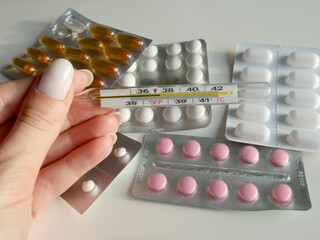 A close-up of a female hand holding a thermometer, surrounded by blister packs of various pills in different colors against a light background. Useful for health and medical-related visuals.